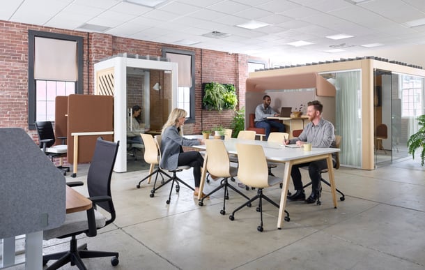 People working in a modern office space with collaborative tables, private work pods, and soft seating areas, surrounded by brick walls and natural greenery.
