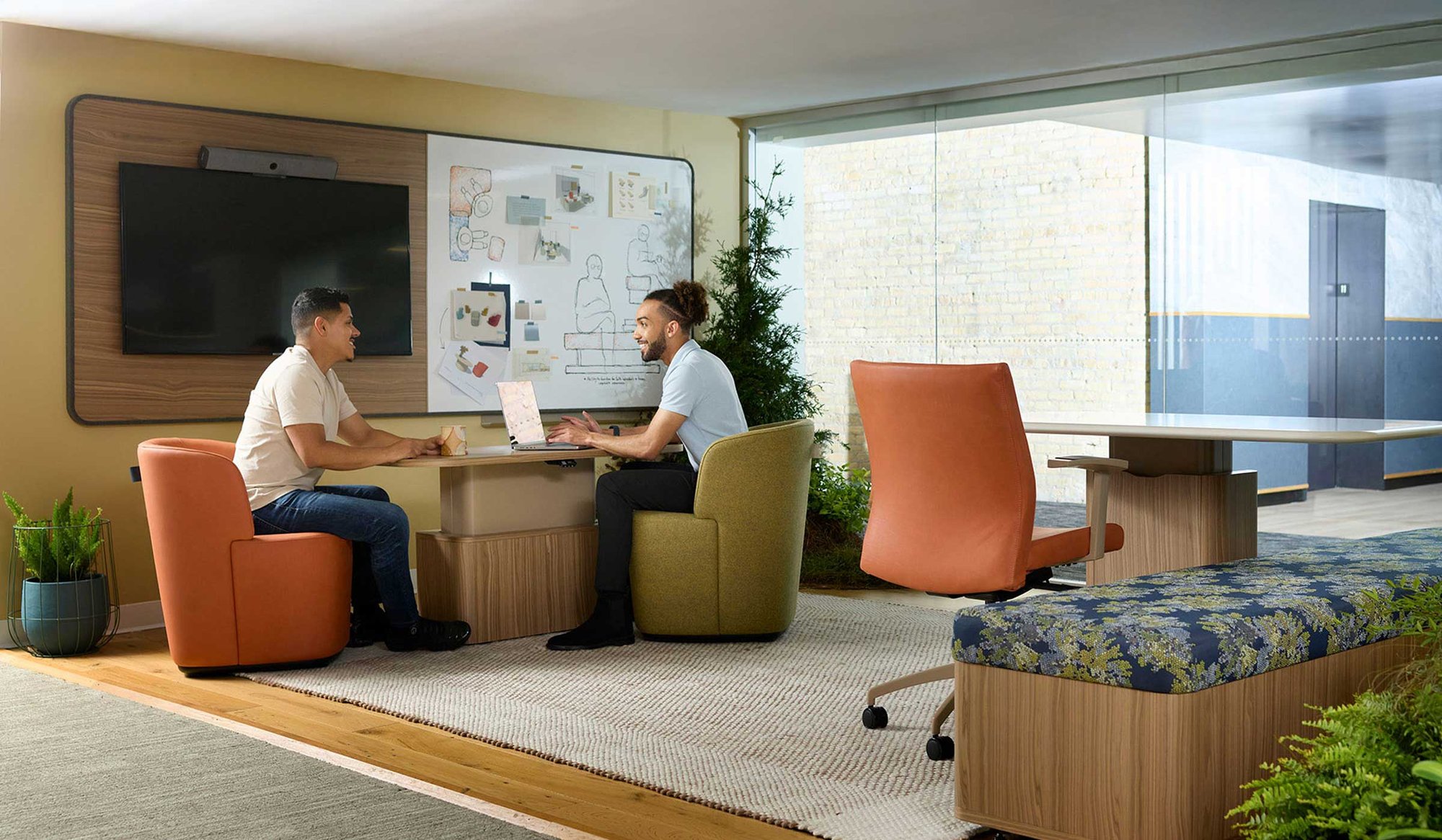 Two colleagues collaborating at a lounge desk with a wall-mounted screen and whiteboard. Two colleagues collaborating at a lounge desk with a wall-mounted screen and whiteboard.