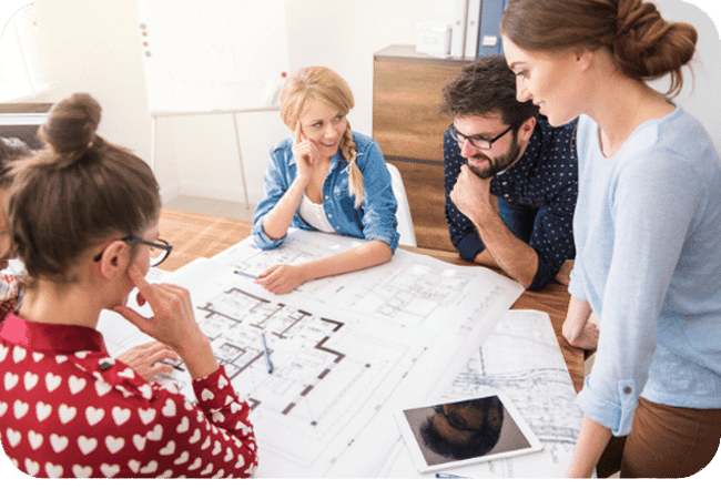 A group of coworkers collaborating around a table, reviewing architectural plans and designs using laptops and digital tablets.