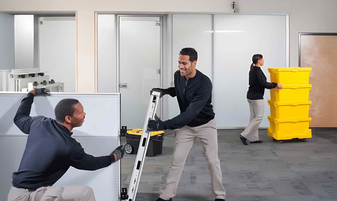 Two workers installing office furniture panels while a colleague moves storage bins.