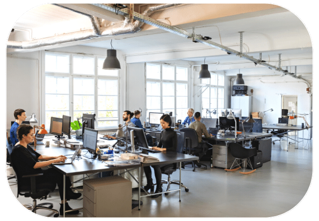 An open office space with multiple people working at desks and computers.